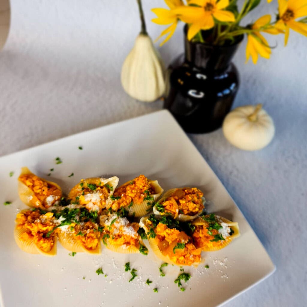 Risotto z mięsem i warzywami podane na białym talerzu, dekoracja pietruszką, obok czarny wazon z żółtymi kwiatami, na szarym tle.