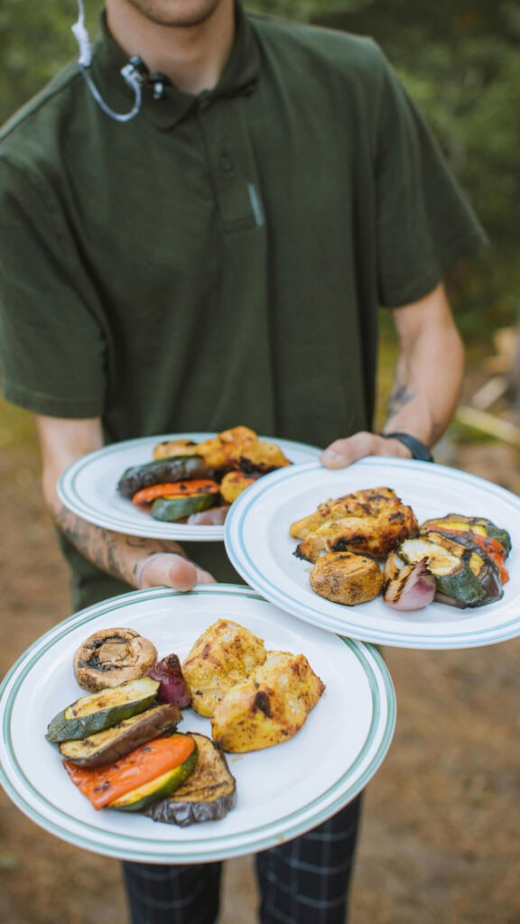 Służba gastronomiczna z talerzami pełnymi grillowanych warzyw i mięs, serwowanymi na świeżym powietrzu w naturalnym otoczeniu.