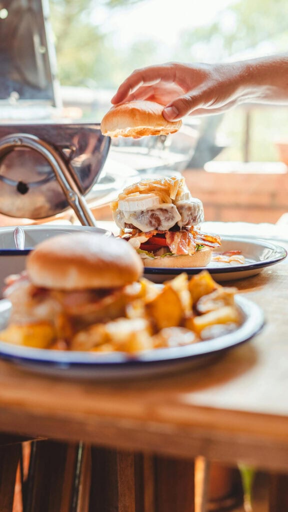 Pracująca ręka układająca bułkę na hamburgerze z mięsem oraz frytkami na tle naturalnego, słonecznego światła.