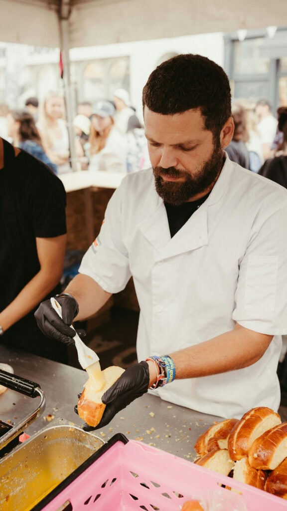 Pracownik przygotowuje kanapkę w strefie gastronomicznej podczas wydarzenia plenerowego. Tło pełne ludzi i stoiska z jedzeniem.