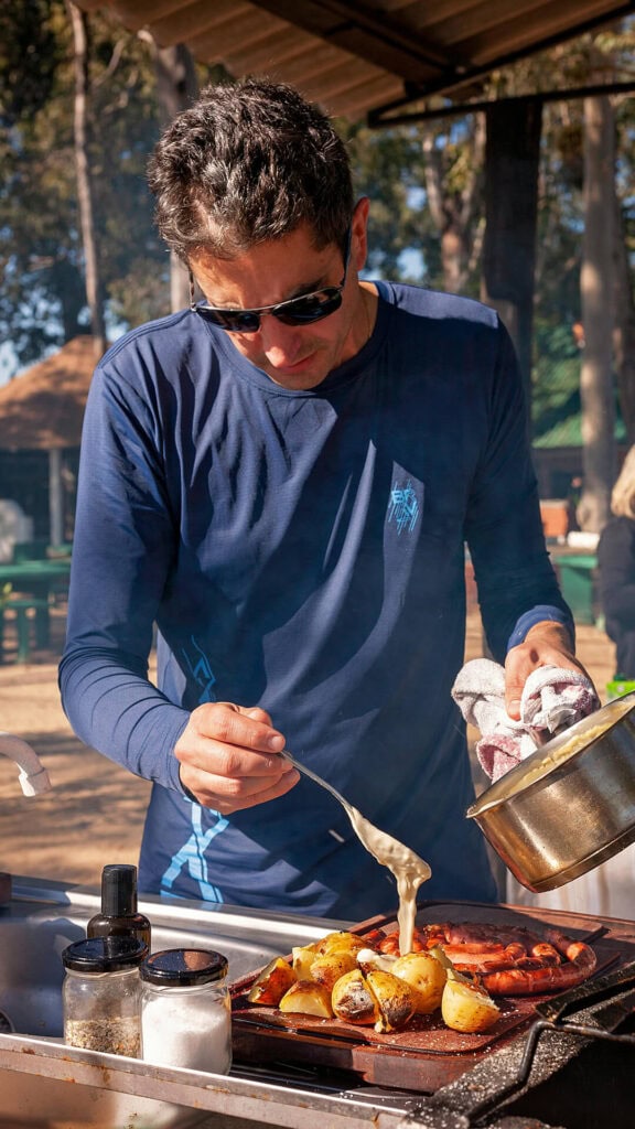 Jest to zdjęcie mężczyzny w ciemnoniebieskiej koszulce, smażącego jedzenie na grillowy sposób w plenerze, wśród drzew i naturalnego otoczenia.