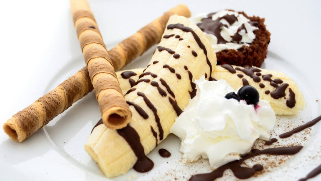 Banana ice cream with chocolate drizzle, whipped cream, and two rolled wafer cookies on a white plate.