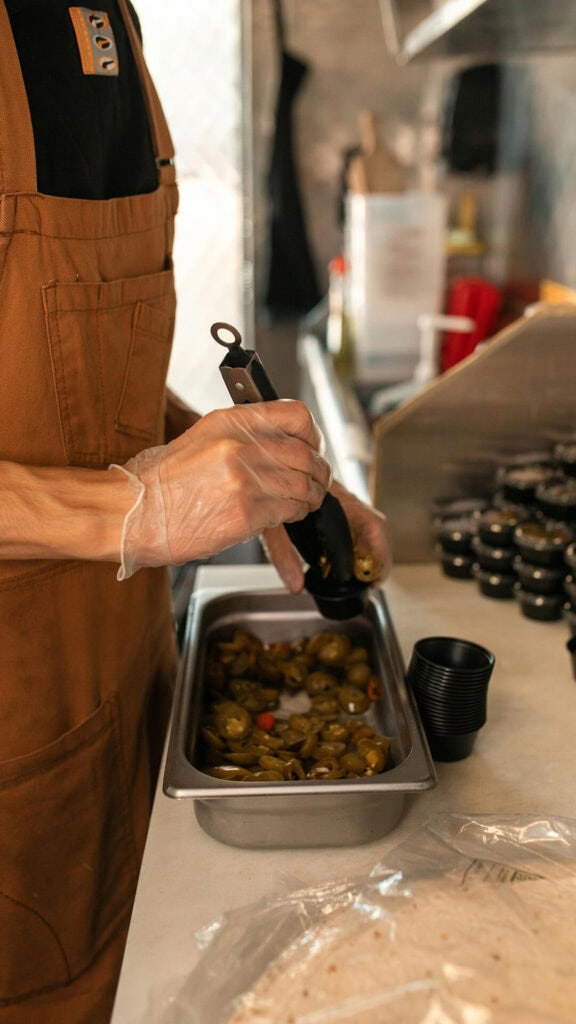 Szczegółowa fotografia osoby w jednolitej odzieży w trakcie przygotowywania potraw w profesjonalnej kuchni, z naciskiem na ręce i składniki.